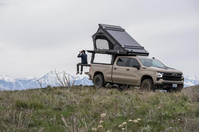 Chevrolet Silverado pickup truck with Lone Peak camper sits on a grassy hill overlooking a mountain range with a man sitting on the edge of a TruckGlide sliding cargo platform extending from the truckbed