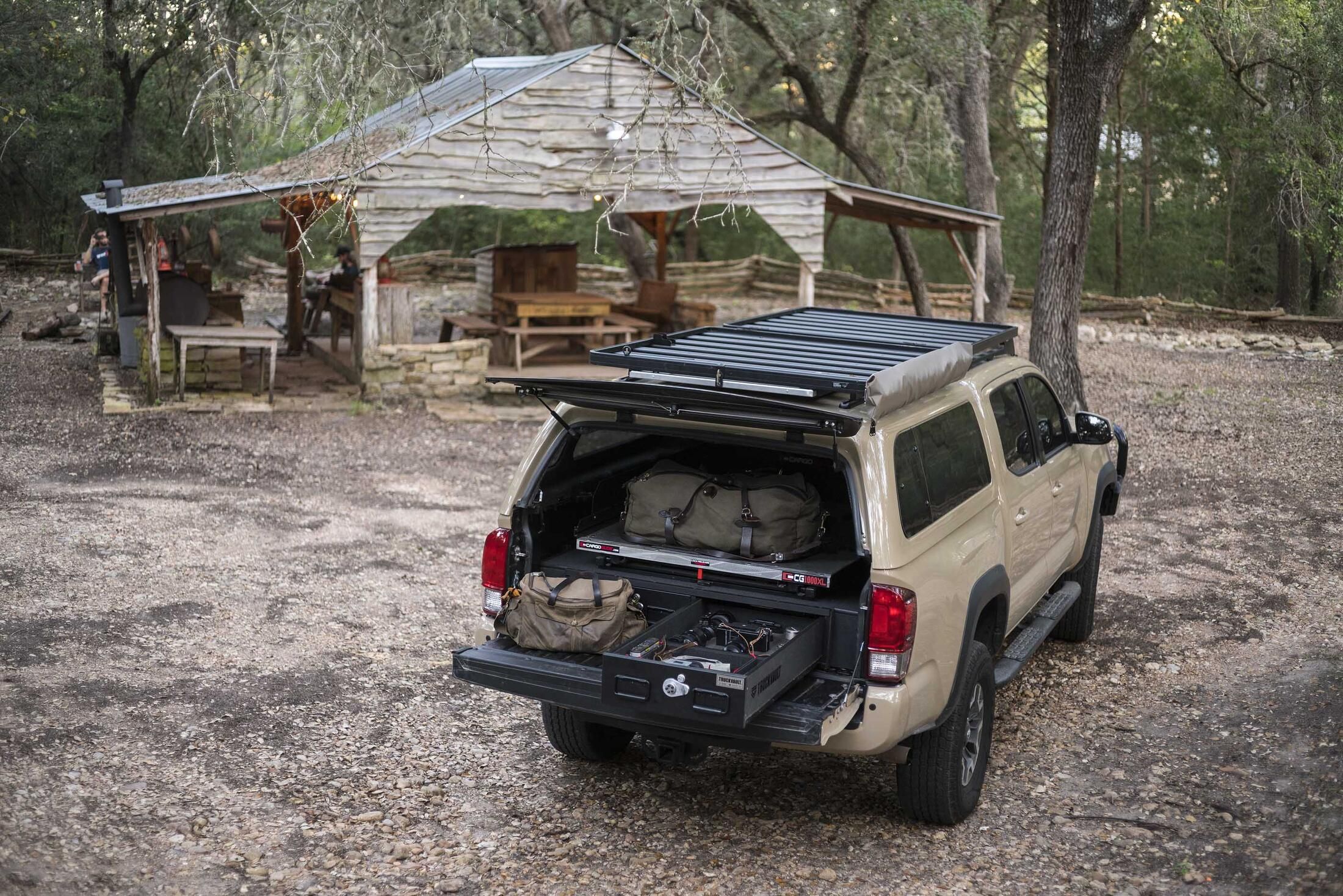 A Toyota Tacoma parked infront of a hut in the woods. Inside of the bed of the Tacoma is an open TruckVault filled with gear.