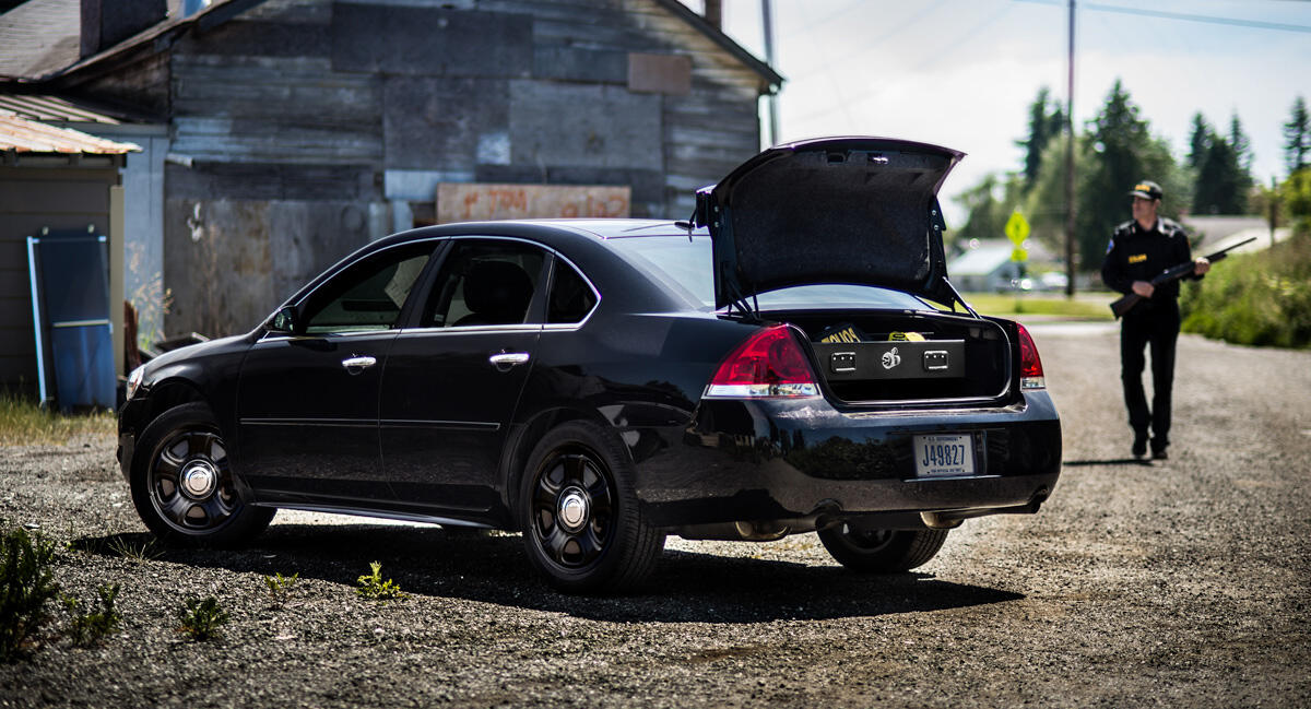 A police officer with a shotgun walking to his Impala with a TruckVault in the trunk space.