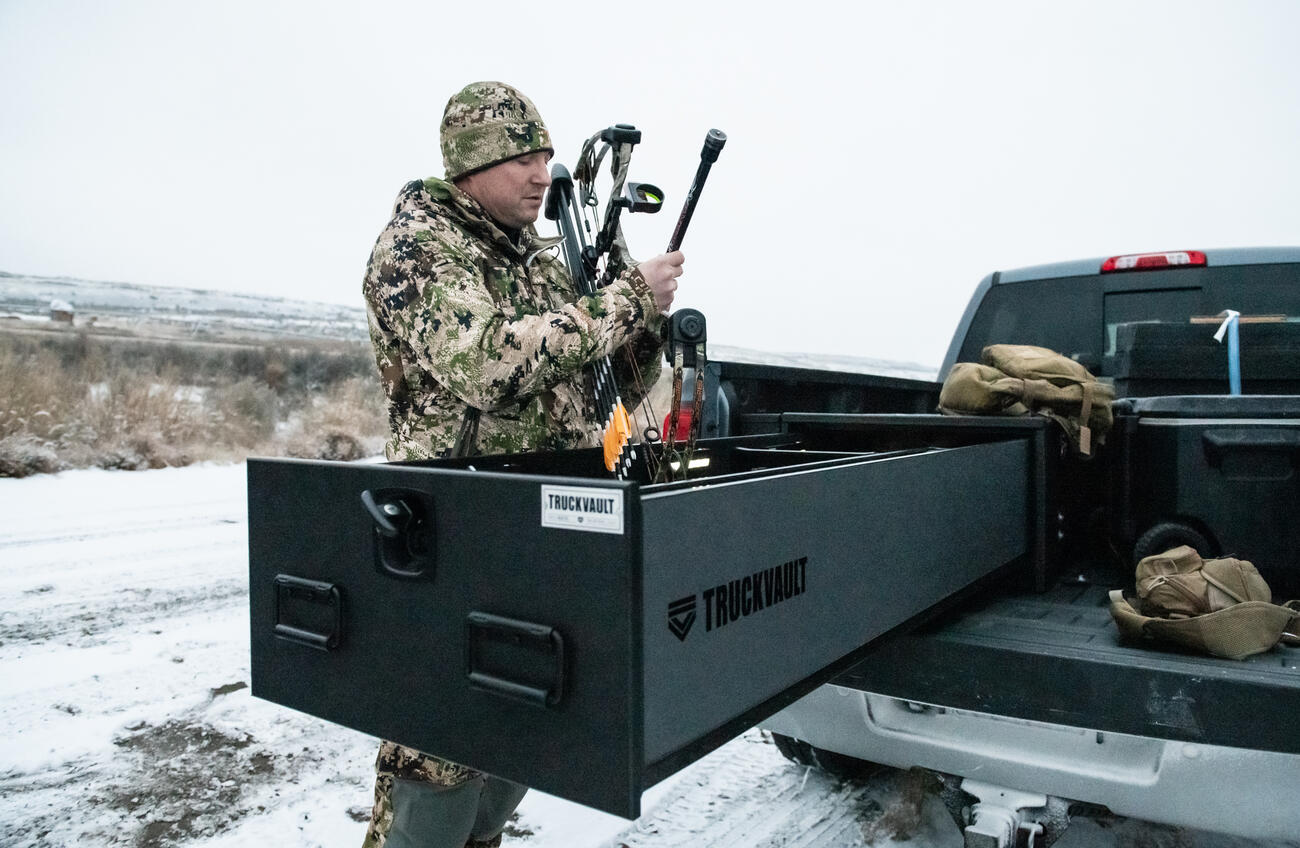 A man in camouflage grabbing his compound bow out of a Half Width TruckVault in the snow.