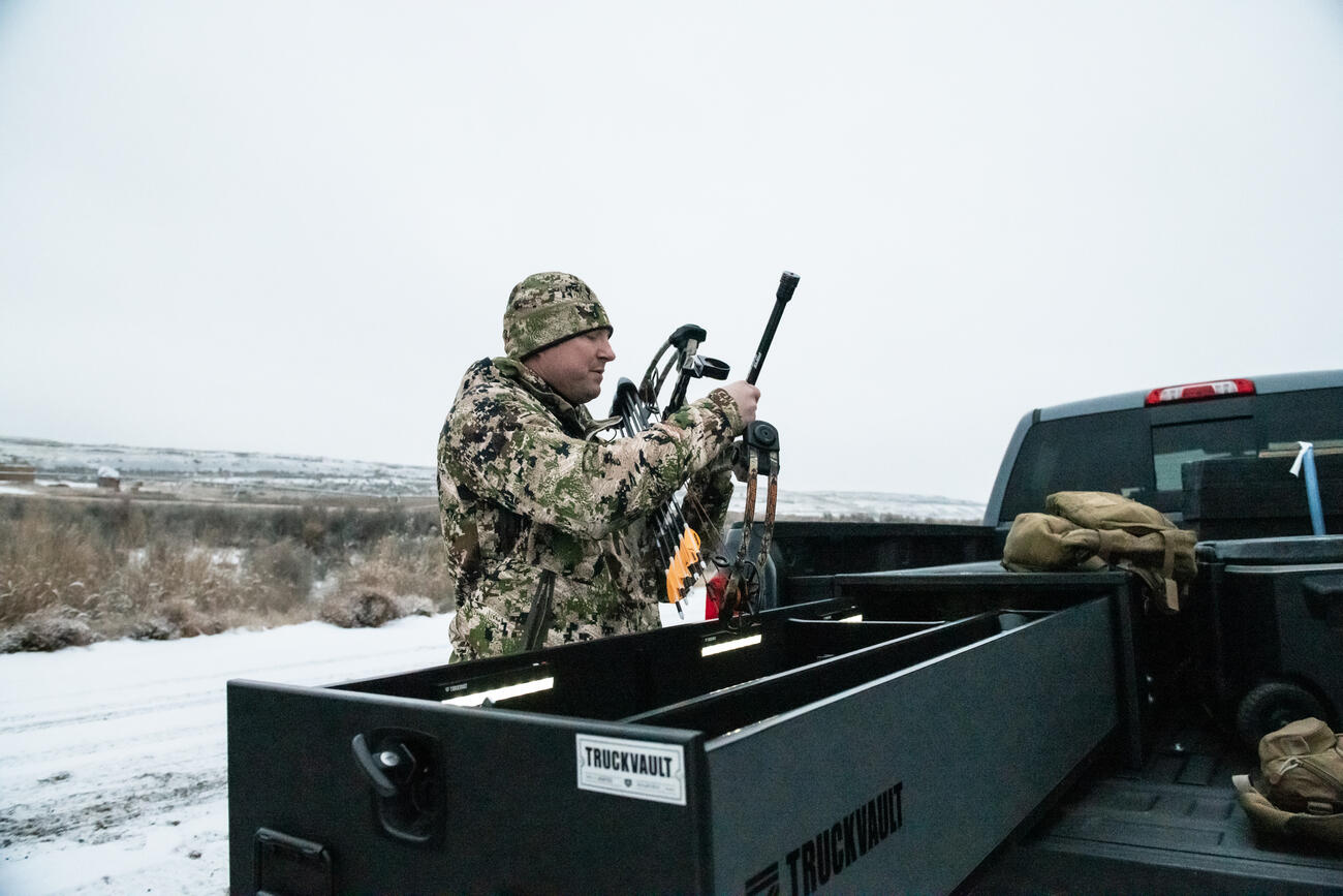 A man in camouflage examining his compound bow near a Half Width TruckVault in the snow.