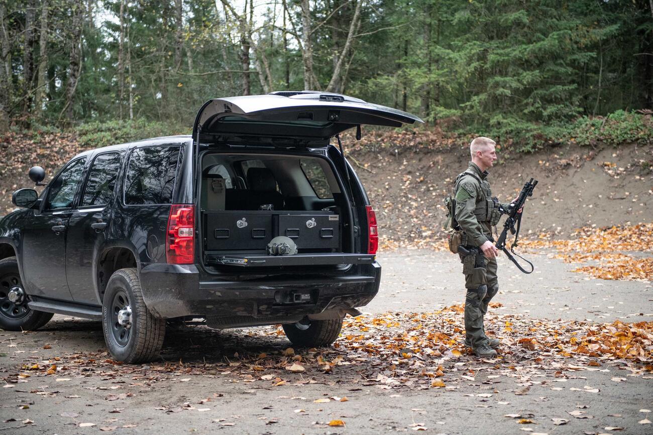 A man in tactical clothing holding a gun walking behind a black Chevy Tahoe complete with a TruckVault and a heavy duty pull out table.