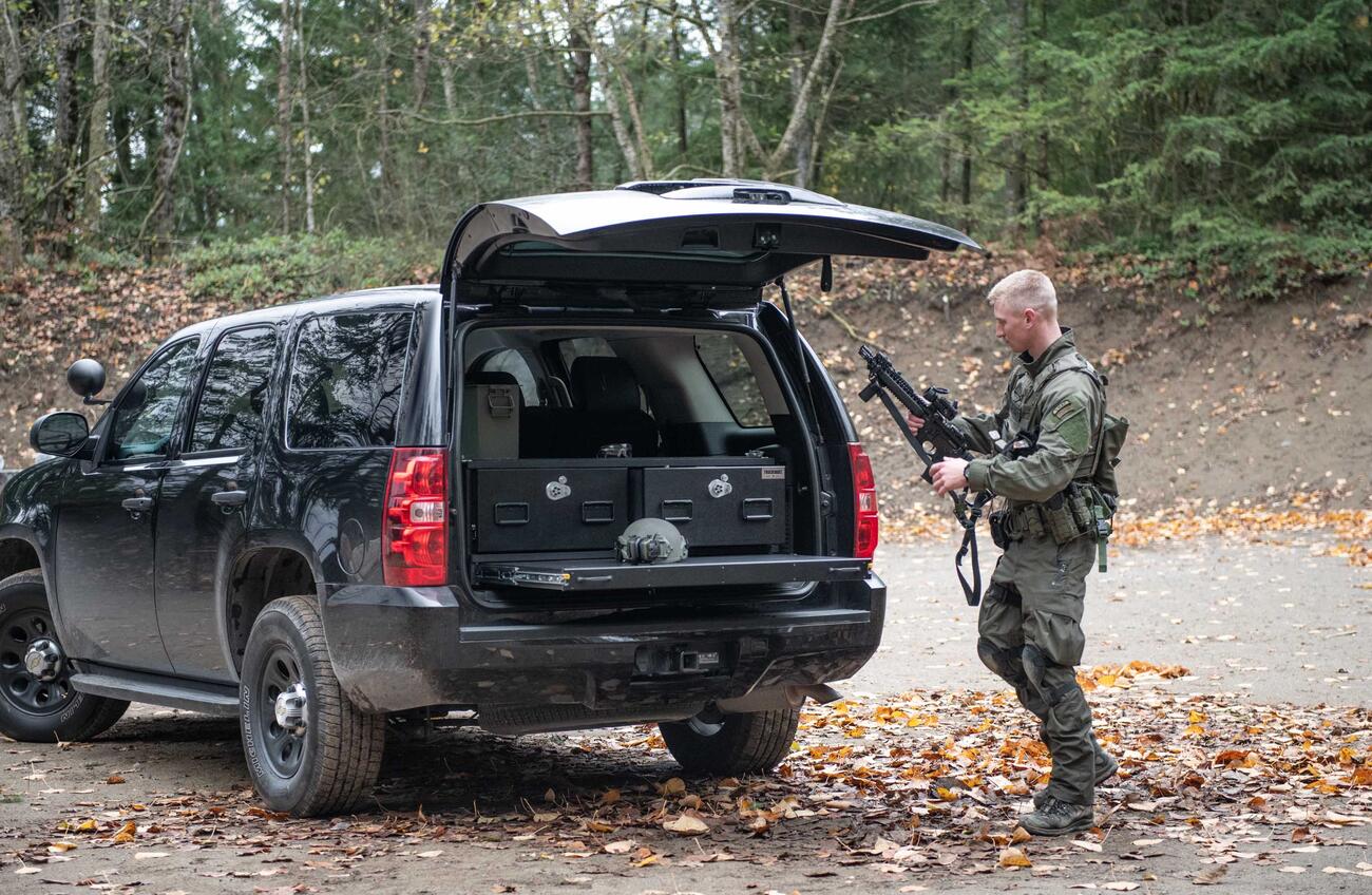 A man in tactical clothing holding a gun walking behind a black Chevy Tahoe complete with a TruckVault and heavy duty pull out table.