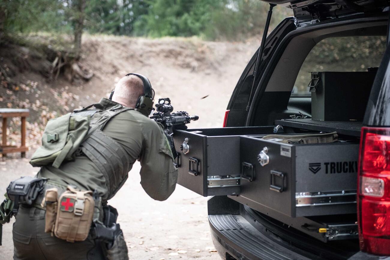 A man shooting a gun that is resting on a TruckVault in the back of a black Chevy Tahoe.