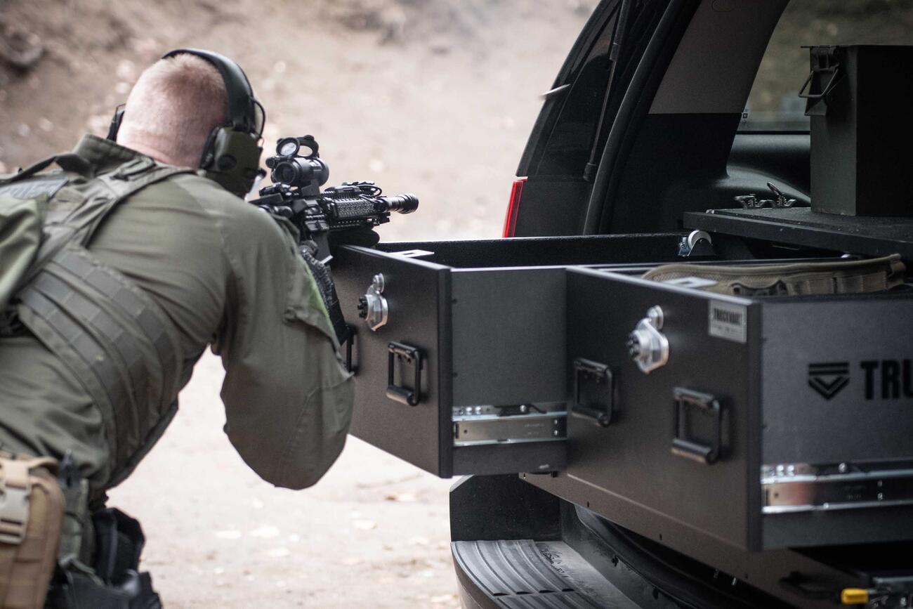 A man shooting a gun that is resting on a TruckVault in the back of a black Chevy Tahoe.