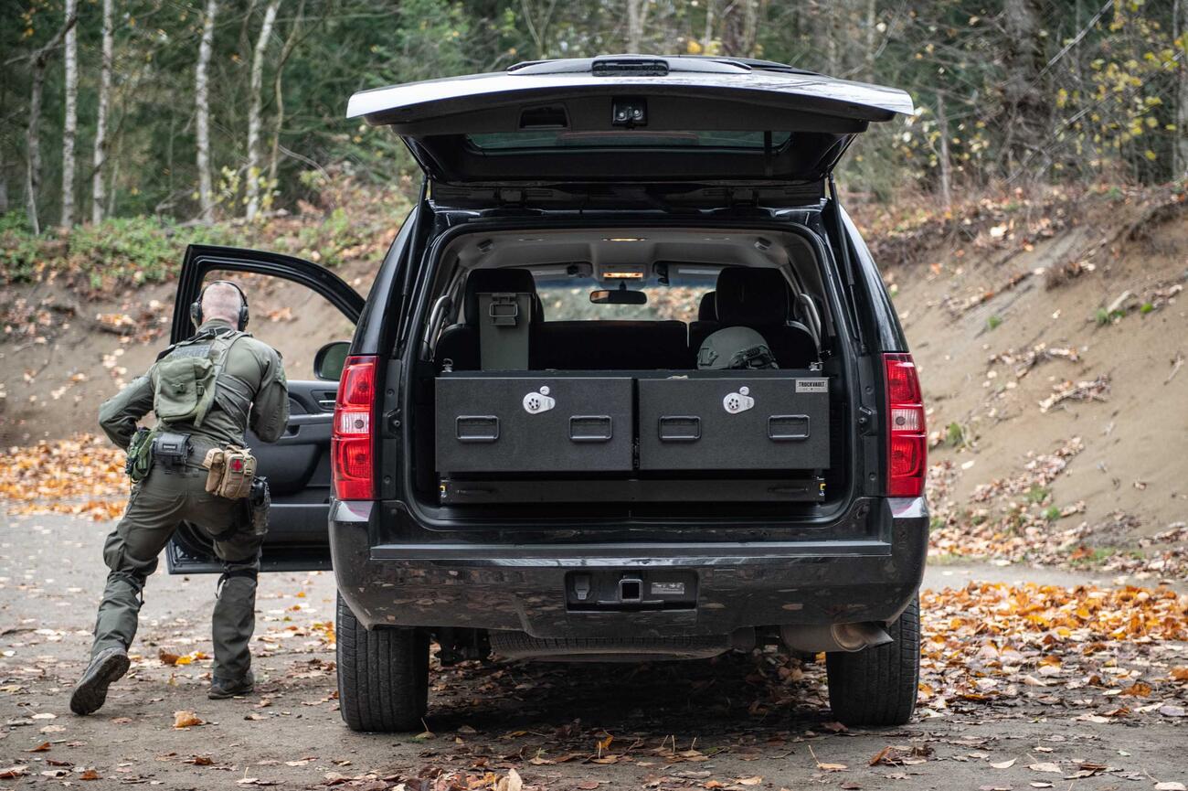 A man in tactical clothing taking cover behind a black Chevy Tahoe with a TruckVault inside of it.