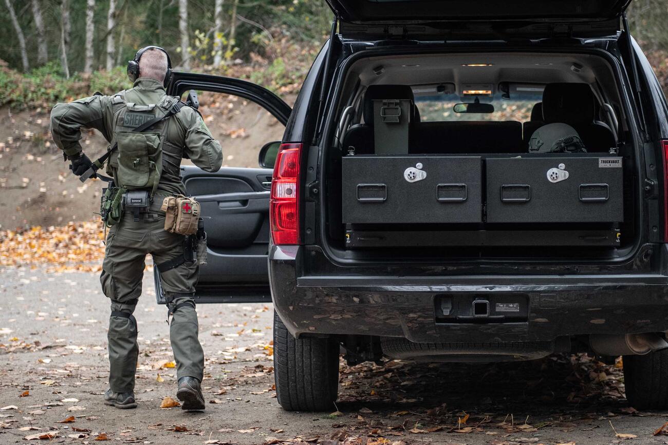 A man with a gun standing next to a black Chevy Tahoe that has a TruckVault in it.