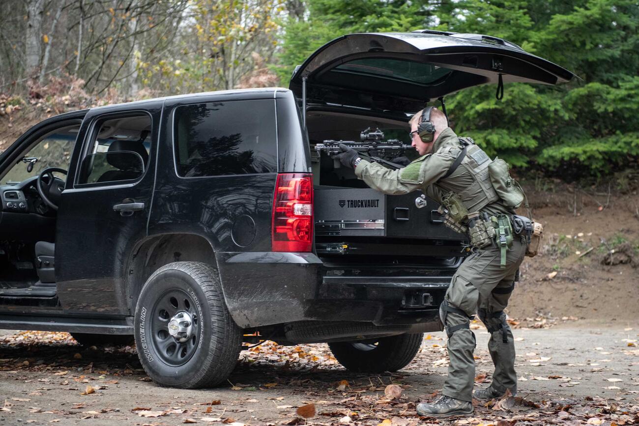 A man shooting a gun that is resting on a TruckVault in the back of a black Chevy Tahoe.