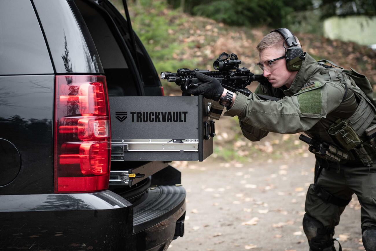 A man Shooting a gun that is resting on a TruckVault in the back of a black Chevy Tahoe.