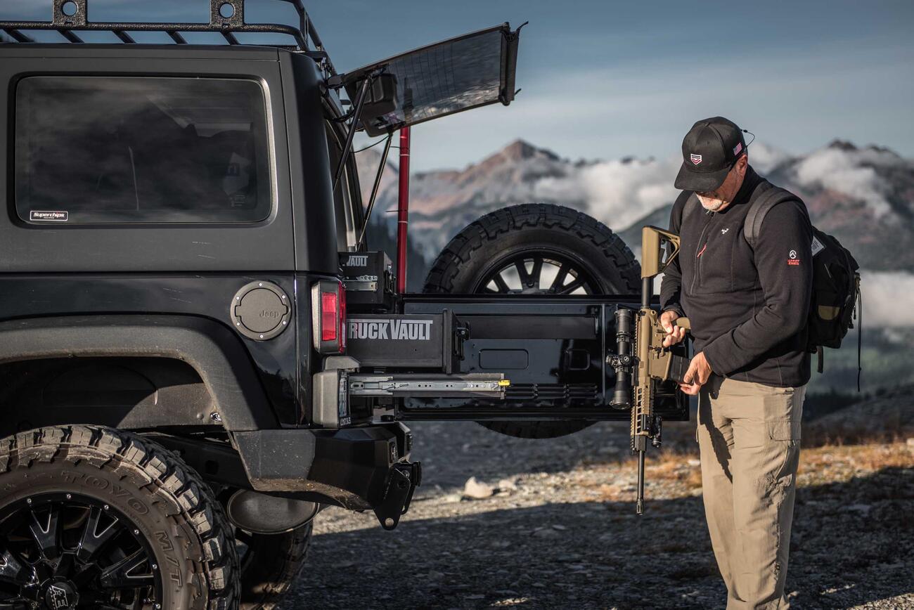 A man holding an assault rifle behind a black Jeep Wrangler with custom TruckVault in the mountains.