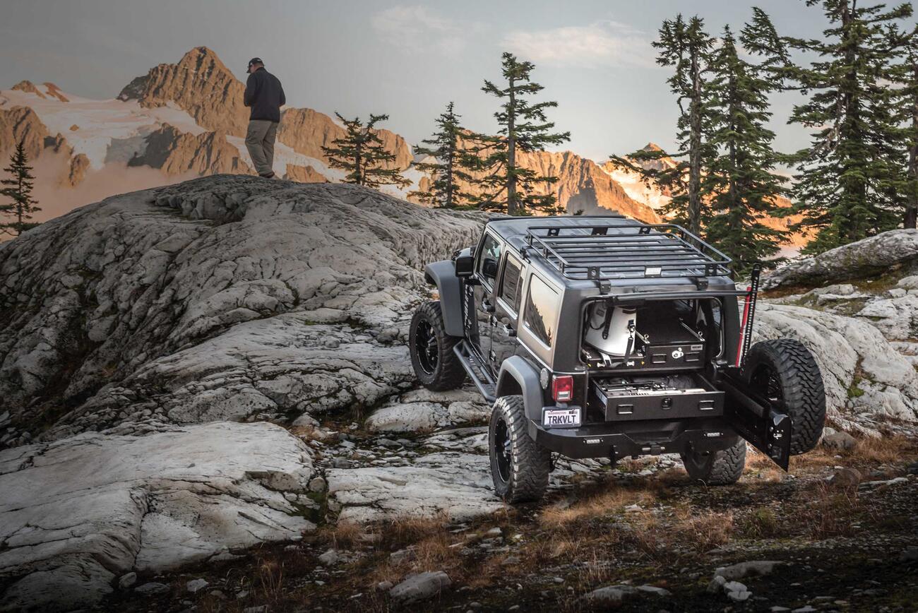 A gray Jeep Wrangler with a custom TruckVault parked on rocks in a mountain range with a man overlooking a valley.