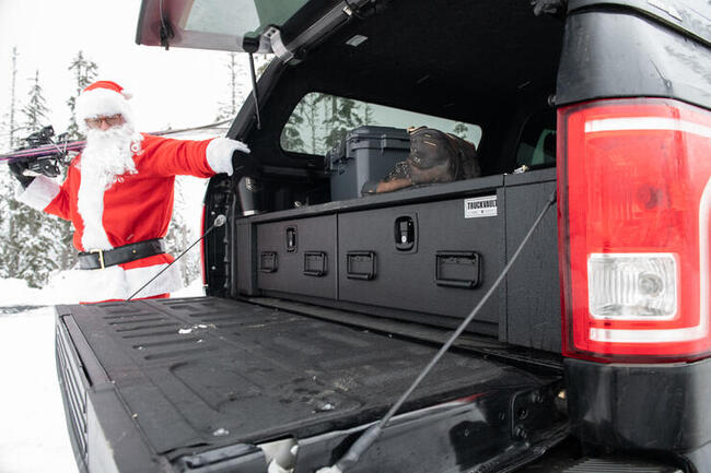 Santa carries skis and reaches into an open truck bed displaying an All Weather TruckVault drawer system