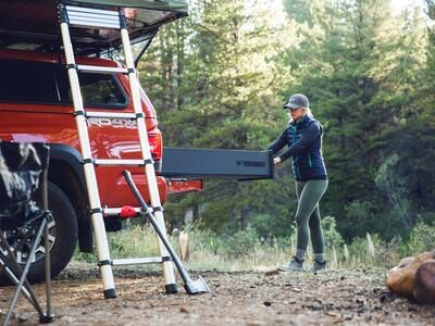 A woman opening a TruckVault in the back of a red truck, in the woods.