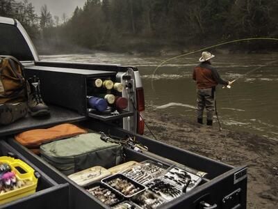 A Silver Ford F-150 filled with fishing gear and a man casting a line in the background.