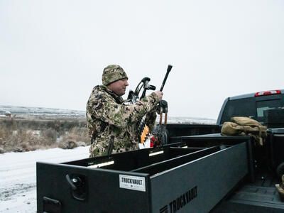 A man in camouflage examining his compound bow near a Half Width TruckVault in the snow.