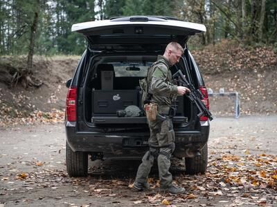 A man in tactical clothing holding a gun walking behind a black Chevy Tahoe complete with a TruckVault and a heavy duty pull out table.