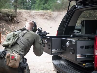 A man shooting a gun that is resting on a TruckVault in the back of a black Chevy Tahoe.