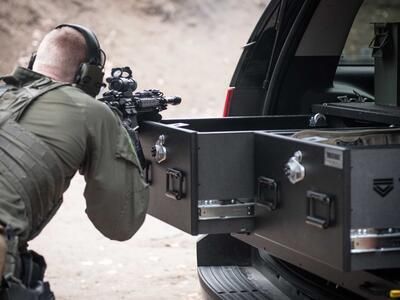 A man shooting a gun that is resting on a TruckVault in the back of a black Chevy Tahoe.