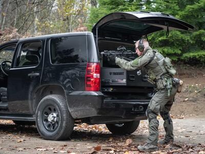 A man shooting a gun that is resting on a TruckVault in the back of a black Chevy Tahoe.