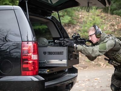 A man Shooting a gun that is resting on a TruckVault in the back of a black Chevy Tahoe.