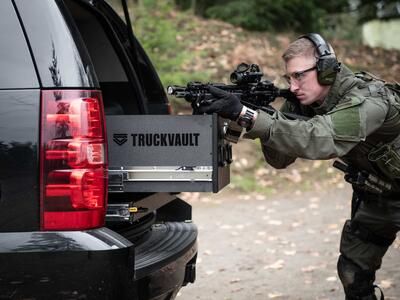 A man Shooting a gun that is resting on a TruckVault in the back of a black Chevy Tahoe.