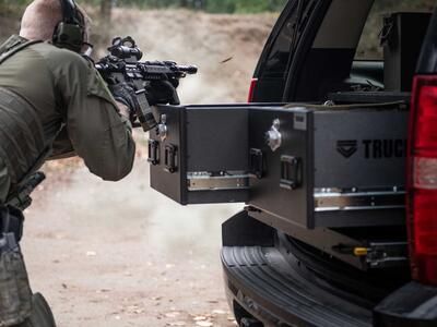 A man shooting a gun that is resting on a TruckVault in the back of a black Chevy Tahoe.