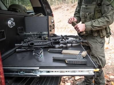 A man is loading his magazine while his rifle rests on a heavy duty pull out table in the back of a black Chevy Tahoe